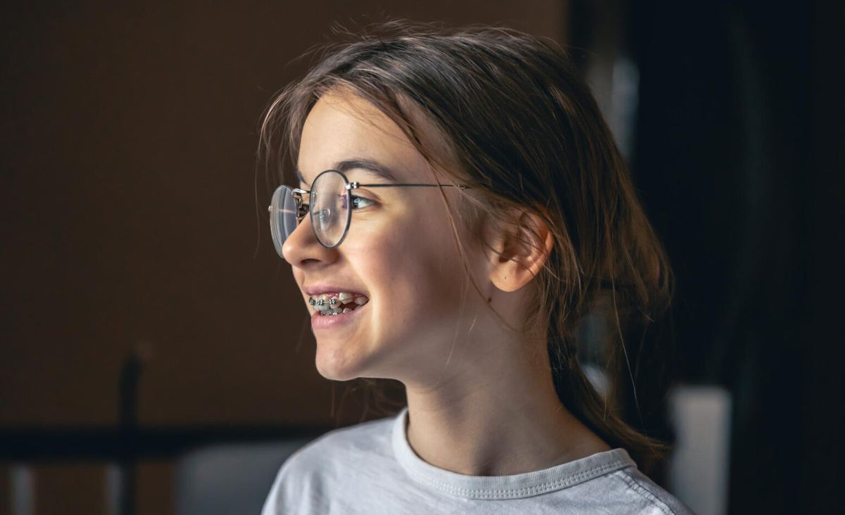 Young girl with braces wearing glasses, smiling and looking away, showing off her orthodontic treatment progress