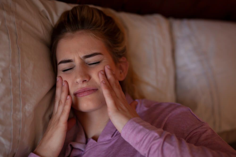 Woman in purple shirt lying in bed and rubbing jaw in pain