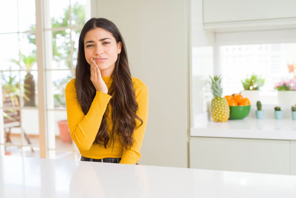 Woman with yellow sweater holding her right hand to her cheek in pain because of a toothache