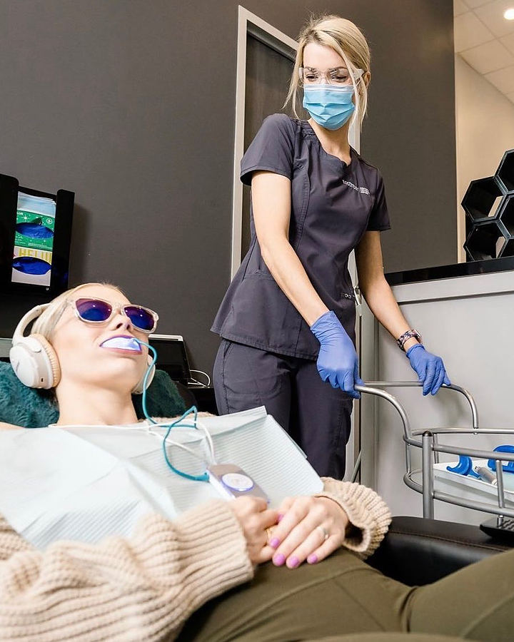 A patient reclined in the chair with protective glasses and headphones during in-office whitening, next to a hygienist