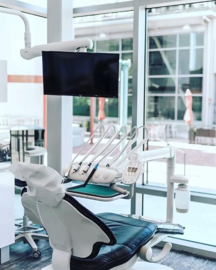 A treatment suite with a black dental chair, a ceiling-mounted screen and tall windows